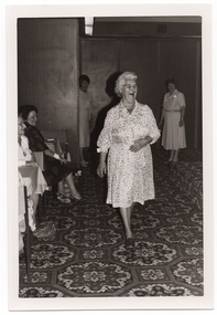 B&W photo of a woman, smiling broadly, modelling a three-quarter sleeve spotted dress in a light colour. She is walking across the bold-patterned carpeted floor below a low stage (which is covered in the same carpet). In the background, there are two other women - one is standing partly behind a wooden partition and the other is standing on the floor beside the low stage. A few members of the seated audience are evident to the left of the frame. There are two or more bags on the floor, at their feet.
