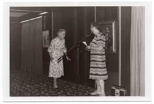 B&W photo of two women on a low stage. One of the women, who is modelling a long-sleeved patterned dress in a light colour, is approaching the other woman, who is standing behind a microphone on a stand. She is wearing a light and dark patterned short-sleeved dress and holding a couple of pieces of paper in her hands. There are two framed pictures on the wall at the back of the stage, and a curtained section at either end. There is a portable radio/cassette player on the floor behind the woman at the microphone. There is a bold patterned carpet on the stage floor.