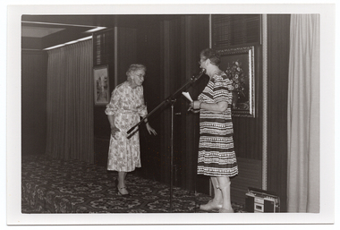 B&W photo of two women on a low stage. One of the women, who is modelling a long-sleeved patterned dress in a light colour, is approaching the other woman, who is standing behind a microphone on a stand. She is wearing a light and dark patterned short-sleeved dress and holding a couple of pieces of paper in her hands. There are two framed pictures on the wall at the back of the stage, and a curtained section at either end. There is a portable radio/cassette player on the floor behind the woman at the microphone. There is a bold patterned carpet on the stage floor.
