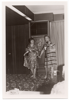 B&W photo of two women on a low stage. One of the women is modelling a short-sleeved dress with a leaf pattern in a medium tone. She is pinching the skirt of her dress between her fingers and lifting it up slightly. She is holding a walking stick in her other hand. The other woman is standing behind a microphone on a stand. She is wearing a light and dark short-sleeved patterned dress. She is holding two pieces of paper in one hand and has one finger of her other hand hooked around the finger of the woman holding the walking stick. Behind them, there is a framed picture of a floral arrangement on the wall. There is a bold patterned carpet on the stage floor.
