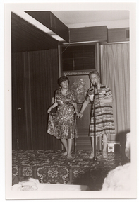 B&W photo of two women on a low stage. One of the women is modelling a short-sleeved dress with a leaf pattern in a medium tone. She is pinching the skirt of her dress between her fingers and lifting it up slightly. She is holding a walking stick in her other hand. The other woman is standing behind a microphone on a stand. She is wearing a light and dark short-sleeved patterned dress. She is holding two pieces of paper in one hand and has one finger of her other hand hooked around the finger of the woman holding the walking stick. Behind them, there is a framed picture of a floral arrangement on the wall. There is a bold patterned carpet on the stage floor.