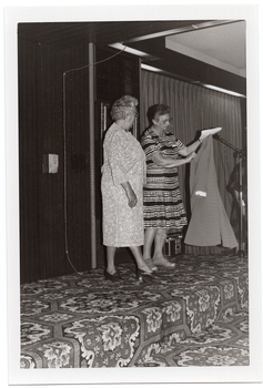 B&W photo of three women on a low stage. One woman is modelling a three-quarter sleeved dress with a flower pattern in a medium tone. She is standing next to another woman who is holding up a coat in a medium tone. She is wearing a light and dark patterned and striped short-sleeved dress. To the right of the frame, there is a microphone on a stand, behind which third woman (further along the stage and partially obscured) is evident. There is a bold patterned carpet on the stage floor.