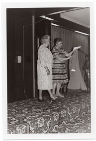 B&W photo of three women on a low stage. One woman is modelling a three-quarter sleeved dress with a flower pattern in a medium tone. She is standing next to another woman who is holding up a coat in a medium tone. She is wearing a light and dark patterned and striped short-sleeved dress. To the right of the frame, there is a microphone on a stand, behind which third woman (further along the stage and partially obscured) is evident. There is a bold patterned carpet on the stage floor.