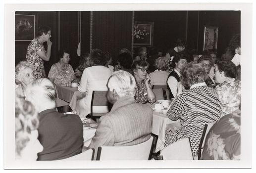B&W photo of a big group of people seated at several rows of tables covered with tablecloths. On top of the tables, there are cups and saucers, glasses, salt and pepper shakers, and small plates. There are three people standing in between the rows of tables. There are three framed pictures on the wall.