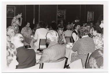 B&W photo of a big group of people seated at several rows of tables covered with tablecloths. On top of the tables, there are cups and saucers, glasses, salt and pepper shakers, and small plates. There are three people standing in between the rows of tables. There are three framed pictures on the wall.