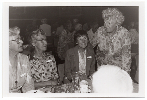 B&W medium close up photo of four women at a table. Three of the women are seated at the table, and a fourth woman is standing next to the table, leaning forward slightly. On top of the table, there are wine glasses, cups and saucers, small plates, a glass jug, and a glass carafe. In the foreground, to the right of the frame, one person's head has been Photographed from behind. In the background, there are numerous people sitting at other tables.