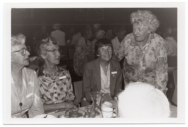 B&W medium close up photo of four women at a table. Three of the women are seated at the table, and a fourth woman is standing next to the table, leaning forward slightly. On top of the table, there are wine glasses, cups and saucers, small plates, a glass jug, and a glass carafe. In the foreground, to the right of the frame, one person's head has been Photographed from behind. In the background, there are numerous people sitting at other tables.