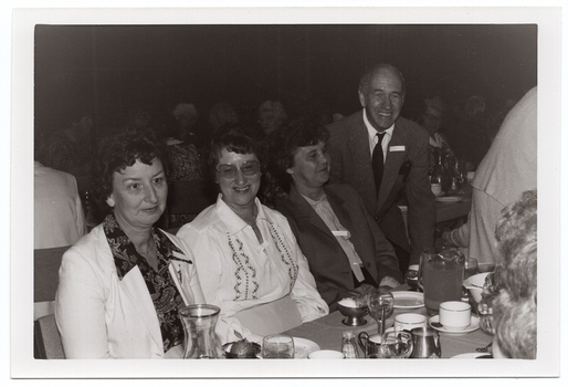 B&W medium close up photo of four people at a table. Three women are seated at the table, and a man is standing next to the table, leaning forward slightly. On top of the table, there are wine glasses, cups and saucers, small plates, a glass jug, stainless steel milk jugs, stainless steel dessert cups, and a glass carafe. In the foreground, to the right of the frame, two other people have been Photographed from behind - one person's head (partially obscured) and a woman leaning forward. In the background, there are numerous people sitting at other tables.