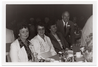 B&W medium close up photo of four people at a table. Three women are seated at the table, and a man is standing next to the table, leaning forward slightly. On top of the table, there are wine glasses, cups and saucers, small plates, a glass jug, stainless steel milk jugs, stainless steel dessert cups, and a glass carafe. In the foreground, to the right of the frame, two other people have been Photographed from behind - one person's head (partially obscured) and a woman leaning forward. In the background, there are numerous people sitting at other tables.
