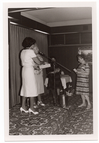 B&W photo of four women on or near a low stage. Two of the women are standing behind a microphone on a stand, which is on the stage. One of them is wearing a pale coloured shirt and skirt, and holding a small box. The other woman is wearing a patterned top and striped skirt, and speaking into the microphone. A third woman is standing in the background, near one end of the stage. She is wearing a dark coloured patterned dress. Behind her, there is a small table covered with a white tablecloth, on top of which there are a few miscellaneous items. In front of the table, on the bold-patterned carpeted stage floor, there is a gift basket decorated with ribbons. There is a fourth woman standing on the floor below the low stage. She is wearing a light and dark patterned short-sleeved dress. She is holding some pieces of paper in both hands and facing the stage. There is a framed picture on the wall behind her.