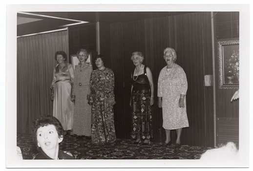 B&W photo of five women standing in a row on a small stage, modelling some clothes. Four of the women are wearing floor-length dresses or skirt (a floor-length skirt and a top with wide straps), and the fifth woman is wearing a three-quarter sleeved, calf length dress. One of the women is using a walking stick. There are two framed pictures on the wall behind them, and a bold patterned carpet on the stage floor. In the foreground, to the left of the frame, a woman in the audience is turning her head to look behind her.