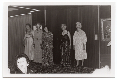 B&W photo of five women standing in a row on a small stage, modelling some clothes. Four of the women are wearing floor-length dresses or skirt (a floor-length skirt and a top with wide straps), and the fifth woman is wearing a three-quarter sleeved, calf length dress. One of the women is using a walking stick. There are two framed pictures on the wall behind them, and a bold patterned carpet on the stage floor. In the foreground, to the left of the frame, a woman in the audience is turning her head to look behind her.