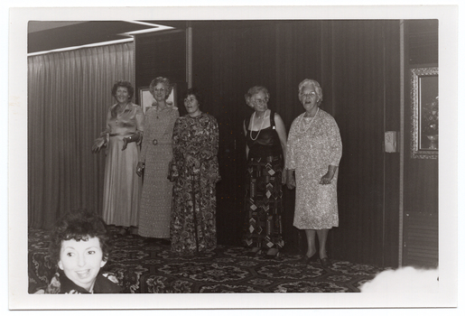 B&W photo of five women standing on a low on a stage, modelling some clothes. Four of the women are wearing floor-length dresses or skirt (a floor-length skirt and a top with wide straps), and the fifth woman is wearing a three-quarter sleeved, calf length dress. One of the women is using a walking stick. There are two framed pictures on the wall behind them, and a bold patterned carpet on the stage floor. In the foreground, to the left of the frame, a woman in the audience is turning her head to look behind her.