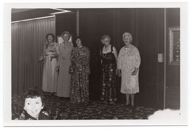 B&W photo of five women standing on a low on a stage, modelling some clothes. Four of the women are wearing floor-length dresses or skirt (a floor-length skirt and a top with wide straps), and the fifth woman is wearing a three-quarter sleeved, calf length dress. One of the women is using a walking stick. There are two framed pictures on the wall behind them, and a bold patterned carpet on the stage floor. In the foreground, to the left of the frame, a woman in the audience is turning her head to look behind her.