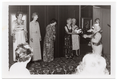B&W photo of six women standing on a low on a stage and a mascot (a person dressed in a rat costumre) is standing on the floor below the stage. The three women standing to the left of the frame are wearing long-sleeved, floor-length dresses. One of them is leaning forward slightly as she smiles and looks towards the women to the right of the frame. She is leaning on a walking stick with both hands. A little further along the stage (to the right of the frame), two women are standing close together. One of them is wearing a floor-length skirt and a top with wide straps, while the other woman is wearing a three-quarter sleeve calf-length dress. The woman in the long skirt is accepting a gift-wrapped potted plant decorated with bows from the mascot. At the far end of the stage, there is a sixth woman (partially obscured by the mascot) standing near a microphone on a stand. There are two framed pictures on the wall at the back of the stage, and a bold patterned carpet on the stage floor. In the foreground, the heads of two audience members have been Photographed from behind.