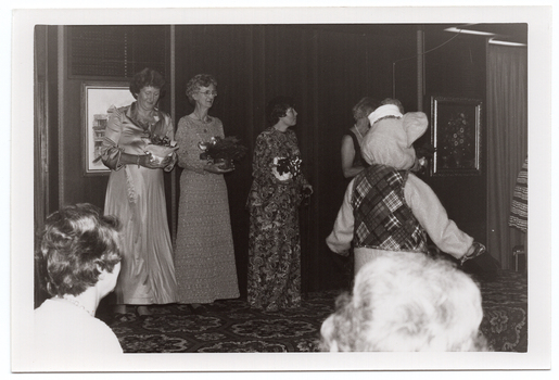 B&W photo of five women on a low stage (two partially obscured), and a mascot (someone in a rat costume) standing on the floor area below the stage. The three women who are not obscured from view are each holding a gift-wrapped potted plant decorated with bows. One of them is looking down at her potted plant. To the far right of the frame, a sixth woman (partially obscured) is standing near the edge of the stage, leaning forward. There are two framed pictures on the wall behind the stage. In the foreground, two audience members' heads have been Photographed from behind.