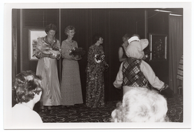 B&W photo of five women on a low stage (two partially obscured), and a mascot (someone in a rat costume) standing on the floor area below the stage. The three women who are not obscured from view are each holding a gift-wrapped potted plant decorated with bows. One of them is looking down at her potted plant. To the far right of the frame, a sixth woman (partially obscured) is standing near the edge of the stage, leaning forward. There are two framed pictures on the wall behind the stage. In the foreground, two audience members' heads have been Photographed from behind.