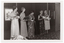 B&W photo of five women standing on a small stage, each holding a gift-wrapped potted plant decorated with bows. One of the women is using a walking stick. She is looking down slightly. Behind them, there are two framed pictures on the wall at the back of the stage. On the right edge of the frame, there is a radio-cassette player (partially obscured) on the bold-patterned carpeted stage floor.