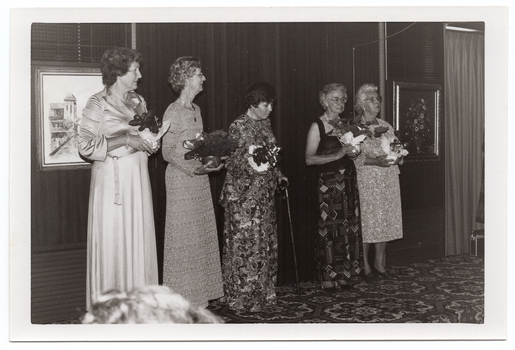 B&W photo of five women standing on a small stage, each holding a gift-wrapped potted plant decorated with bows. One of the women is using a walking stick. She is looking down slightly. Behind them, there are two framed pictures on the wall at the back of the stage. On the right edge of the frame, there is a radio-cassette player (partially obscured) on the bold-patterned carpeted stage floor.