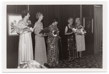 B&W photo of five women standing on a small stage, each holding a gift-wrapped potted plant decorated with bows. One of the women is using a walking stick. She is looking down slightly. Behind them, there are two framed pictures on the wall at the back of the stage. On the right edge of the frame, there is a radio-cassette player (partially obscured) on the bold-patterned carpeted stage floor.