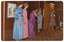 COL photo of seven women on a low stage. Five of the women are standing next to one another in a row. Three of them are wearing floor-length dresses (in blue, pink, and purple) and one is wearing a dark patterned floor-length skirt with a black top with wide straps. The fifth woman is wearing a blue spotted three-quarter sleeve, calf-length dress. The sixth woman is standing further along the stage, speaking into a microphone on a stand. She is wearing a pink, grey, and black patterned striped dress. There is a portable radio/cassette player on the stage floor behind her. At the far end of the stage, there is a seventh woman who is wearing a black dress with a rust or burgundy design throughout. There is a small table with a white tablecloth behind her. There are two framed pictures on the wall behind them, and some curtains along one section behind the stage. The brown carpet on the stage floor has a bold pattern.