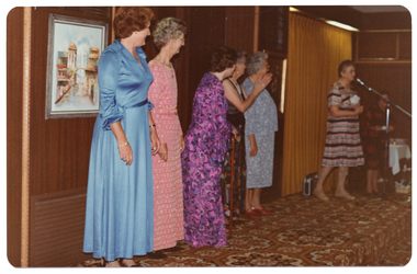 COL photo of seven women on a low stage. Five of the women are standing next to one another in a row. Three of them are wearing floor-length dresses (in blue, pink, and purple) and one is wearing a dark patterned floor-length skirt with a black top with wide straps. The fifth woman is wearing a blue spotted three-quarter sleeve, calf-length dress. The sixth woman is standing further along the stage, speaking into a microphone on a stand. She is wearing a pink, grey, and black patterned striped dress. There is a portable radio/cassette player on the stage floor behind her. At the far end of the stage, there is a seventh woman who is wearing a black dress with a rust or burgundy design throughout. There is a small table with a white tablecloth behind her. There are two framed pictures on the wall behind them, and some curtains along one section behind the stage. The brown carpet on the stage floor has a bold pattern.