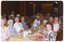 COL photo of a roomful of people sitting at rows of long rectangular tables. There is one group of people sitting at a table in the foreground. On top of the table, there are serving bowls, small and large plates (with cutlery and remnants of a meal), cups and saucers, wine glasses, a glass jug of juice or cordiall, a carafe of white wine, and a glass ashtray. In the background, there are two wood-panelled columns, curtains, and a framed picture on the wall.