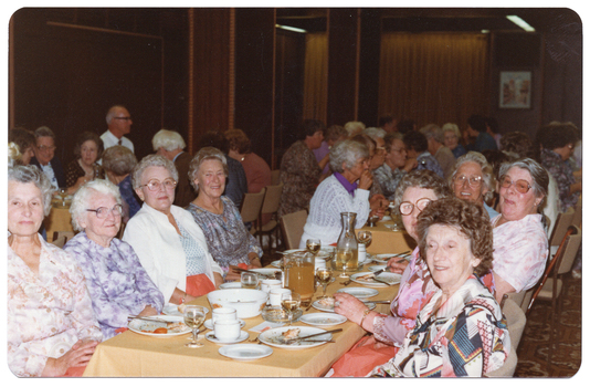 COL photo of a roomful of people sitting at rows of long rectangular tables. There is one group of people sitting at a table in the foreground. On top of the table, there are serving bowls, small and large plates (with cutlery and remnants of a meal), cups and saucers, wine glasses, a glass jug of juice or cordiall, a carafe of white wine, and a glass ashtray. In the background, there are two wood-panelled columns, curtains, and a framed picture on the wall.