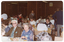 COL photo of a roomful of people sitting at rows of long rectangular tables. Sitting at a table in the foreground, there is a man in a brown suit and a woman in a blue floral dress facing the camera. Sitting at the end of the table behind them, there is a woman with a walking stick leaning against her chair. A few people are standing around the room while chatting to those seated at various tables. In the background, some wood-panelled walls and columns are evident.