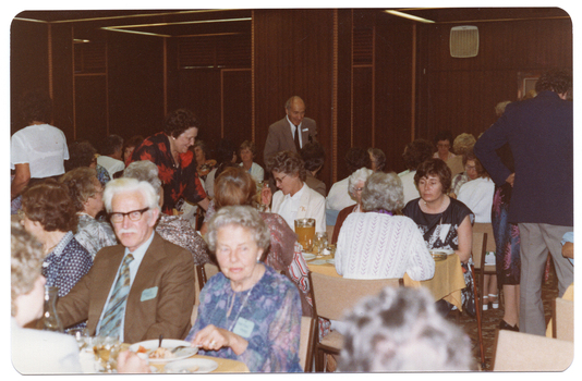 COL photo of a roomful of people sitting at rows of long rectangular tables. Sitting at a table in the foreground, there is a man in a brown suit and a woman in a blue floral dress facing the camera. Sitting at the end of the table behind them, there is a woman with a walking stick leaning against her chair. A few people are standing around the room while chatting to those seated at various tables. In the background, some wood-panelled walls and columns are evident.