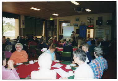 COL wide frame photo of a woman standing in front of a roomful of people, seated at tables. She is speaking into a microphone. There is a wooden lectern beside her. Behind her, there is a slide image being projected onto a freestanding screen and a tall, freestanding blue banner with gold text and fringing. To the left of the frame, there are some floor to ceiling windows. On the wall behind her, there are various framed pictures and signs. A large seated audience, Photographed from behind, is evident in the foreground.