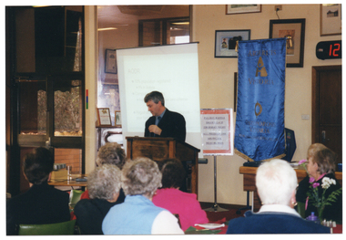 COL photo of a man standing behind a wooden lectern in front of a seated audience. Behind him, there is a projector screen with a slide image being projected onto it. Beside him, on one side, there is a tall, freestanding blue banner with gold text and fringing. On the other side of the room, there are some floor to ceiling windows. On the wall behind him, there are various framed pictures and signs. In the foreground, the seated audience has been Photographed from behind.