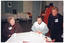 COL photo of a small group of people inside a large room. Two women are seated at an hexagonal table, while another woman is standing next to them. On the table, there are cups and saucers and two cakes on a small plate. A few other people (partially obscured) are standing behind them, to the right of the frame. In the background, there is a long rectangular table covered with a white tablecloth against the wall. On top of the table, there is a large urn, cups and saucers, carafes of water, and glasses. Beneath the table, there is a small waste bin. Next to the table, there is a chair with a tray leaning on the arm.