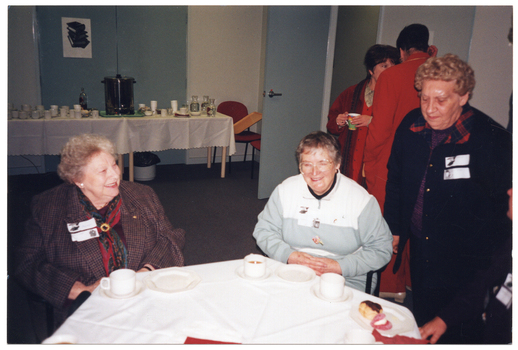 COL photo of a small group of people inside a large room. Two women are seated at an hexagonal table, while another woman is standing next to them. On the table, there are cups and saucers and two cakes on a small plate. A few other people (partially obscured) are standing behind them, to the right of the frame. In the background, there is a long rectangular table covered with a white tablecloth against the wall. On top of the table, there is a large urn, cups and saucers, carafes of water, and glasses. Beneath the table, there is a small waste bin. Next to the table, there is a chair with a tray leaning on the arm.