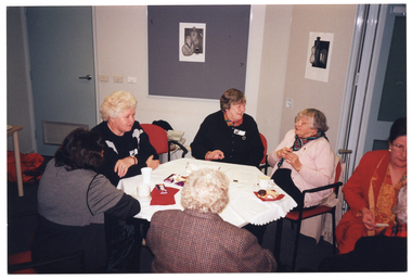 COL photo of five women sitting at an hexagonal table, with two other women (one partially obscured) sitting on chairs opposite one another, next to the table. On the table, there are cups and saucers, a tumbler, a few small blocks of chocolate, and some cakes on a small plate. There are also a couple of pieces of printed paper on the table.
