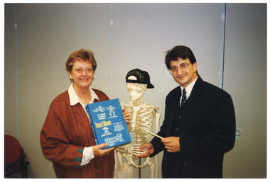 COL photo of a woman and a man posing for a photo with a plastic skeleton, which is on a vertical stand between them. The woman is holding a book or folder titled, "Smart Moves". The man is using a plastic pointing stick to point to the book while he shakes hands with the skeleton. The skeleton is wearing a black cap, back to front, which has the words "SMART MOVES" printed in gold across the back.