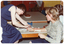 COL photo of three people (one partially obscured) sitting around a low table. One woman (seated) has one hand resting on the table while the other woman (sitting on the table) is fitting a splint onto her hand, wrist and forearm. Only the bottom half of the splint has been fitted, and there are several other pre-cut pieces on the table. There is a chocolate cake on an adjoining table behind.