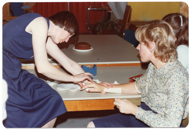COL photo of three people (one partially obscured) sitting around a low table. One woman (seated) has one hand resting on the table while the other woman (sitting on the table) is fitting a splint onto her hand, wrist and forearm. Only the bottom half of the splint has been fitted, and there are several other pre-cut pieces on the table. There is a chocolate cake on an adjoining table behind.