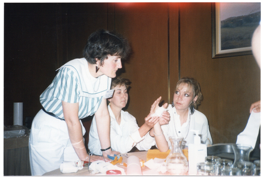 COL photo of a few people gathered around a table. Two women are sitting and two are standing (one predominently obscured). One seated woman is holding her arm up, with her elbow resting on the table. Her hand has a bandage around the palm and thumb. The other seated woman is wrapping the bandage around her hand. Another woman (standing) is leaning forward, with both hands on the table. On the right edge of the frame, another person (predominently obscured) is holding part of a splint in her hand. There are glasses and carafes of water on the table, as well as a pair of scissors, a pair of pliers, some wire, and various unidentified items.