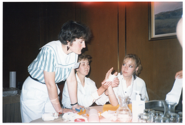 COL photo of a few people gathered around a table. Two women are sitting and two are standing (one predominently obscured). One seated woman is holding her arm up, with her elbow resting on the table. Her hand has a bandage around the palm and thumb. The other seated woman is wrapping the bandage around her hand. Another woman (standing) is leaning forward, with both hands on the table. On the right edge of the frame, another person (predominently obscured) is holding part of a splint in her hand. There are glasses and carafes of water on the table, as well as a pair of scissors, a pair of pliers, some wire, and various unidentified items.