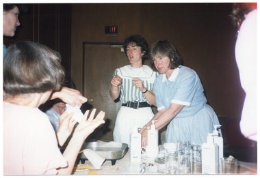 COL photo of a small group of people gathered around a table. Most of the women are standing, while one person is sitting down. One woman is pointing to two women who are holding up a section of fabric in their hands. There are various items on the table, including an electric frying pan, several plastic bottles with pump dispensers, some metal scissors, drinking glasses, paper and other miscellaneous items.