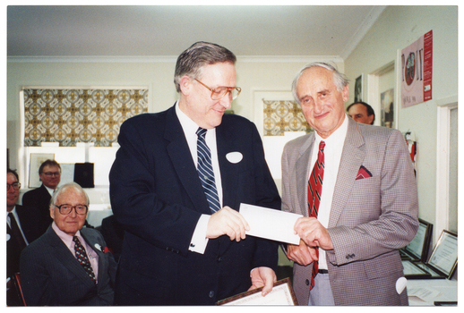 COL photo of several men in a large room. In the foreground, two men are standing next to one another, each holding the edge of a cheque. One of the men is holding a framed certificate in his other hand. There are several framed certificates on a table beside them. Behind them, the other men are either standing or sitting. In the background, there are some large windows with a brown pattened holland blinds and some posters on the wall.