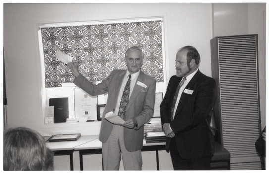 COL photo of two men standing in front of a window with a bold patterned holland blind. One of the men is holding up a cheque in one hand, and an envelope in the other hand. The other man is standing next to him with his arms folded in front of him. He is looking towards the upheld cheque. They are standing in front of two tables beneath a window. On the tables, there are two stacks of framed certificates and other miscellaneous items. On the window sill, there are several items leaning against the window, such as framed certificates, a book, a brochure, and some business cards. In the foreground, to the left of the frame, the back of one person's head is evident. On the right edge of the frame, one person's arms folded over the back of a chair are evident.
