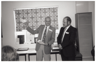 COL photo of two men standing in front of a window with a bold patterned holland blind. One of the men is holding up a cheque in one hand, and an envelope in the other hand. The other man is standing next to him with his arms folded in front of him. He is looking towards the upheld cheque. They are standing in front of two tables beneath a window. On the tables, there are two stacks of framed certificates and other miscellaneous items. On the window sill, there are several items leaning against the window, such as framed certificates, a book, a brochure, and some business cards. In the foreground, to the left of the frame, the back of one person's head is evident. On the right edge of the frame, one person's arms folded over the back of a chair are evident.