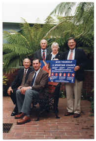 COL photo of five people in a small courtyard with two large ferns and other plants. Two people are sitting on a park bench, and the other three people are standing behind them, holding up a sign titled, "GIVE YOURSELF A SPORTING CHANCE", with the sub-heading, "MANY INJURIES CAN BE PREVENTED". On the sign, there is a photo of someone doing some stretching exercises, some diagrams, and several blocks of text.