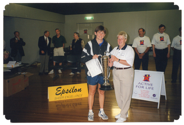 COL photo of two unidentified women posing for a photo while holding a large silver trophy. Behind them, there are several people standing in a row, some of whom are wearing 'Active for Life' branded t-shirts. To the left of the frame, there is a woman sitting at a table and a yellow "Epsilon" sign on the floor. There are boxes underneath the table. To the right of the frame, there is an "Active for Life" A-frame sandwich board on the floor which also contains the logos for the Arthritis Foundation of Victoria and VicHealth.