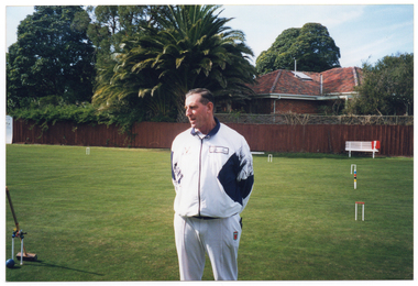 COL photo of a man, wearing a club uniform, standing on a grass croquet court. Hoops are evident at various points on the court, with a centre peg positioned between two hoops. One hoop (on the left edge of the frame) has some croquet clips attached to it and a mallet leaning on it. In the background, there is a white park bench, behind which there is a wooden fence. There is a house and several large trees behind the fence.