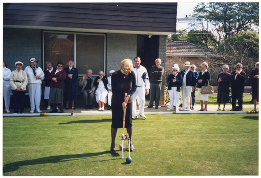 COL photo of a woman on a grass croquet court, standing behind a hoop. She is holding a mallet in position while looking down, and there are several croquet clips attached to the hoop. There is a ball on the other side of the hoop. In the background, there is a group of people gathered alongside a grey brick building, looking on. Almost everyone is laughing, including the woman on the court. Next to the building, behind the fence, there is a house and some trees.