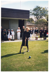 COL photo of a woman on a grass croquet court, standing behind a hoop. She is holding a mallet in position while looking up, and there are several croquet clips attached to the hoop. There is a ball on the other side of the hoop. In the background, there is a group of people gathered alongside a grey brick building, looking on. Next to the building, behind the fence, there is a house and some trees.