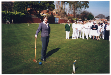 COL photo of a woman, wearing a white shirt, blue trousers and jacket, posing for a photo between two hoops on a grass croquet court. She is holding a mallet, the head of which is resting on the grass. There is a ball near the head of the mallet. Behind her, and to one side, a group of people is gathered on the court. They are wearing white uniforms, a few with either a dark green or dark blue cardigan. In the background, there is a small white bench seat, behind which is a long wooden fence. There is a large sign on the fence, with the text "Bank of Melbourne" along the bottom edge. Behind the fence, there are several houses and large trees.